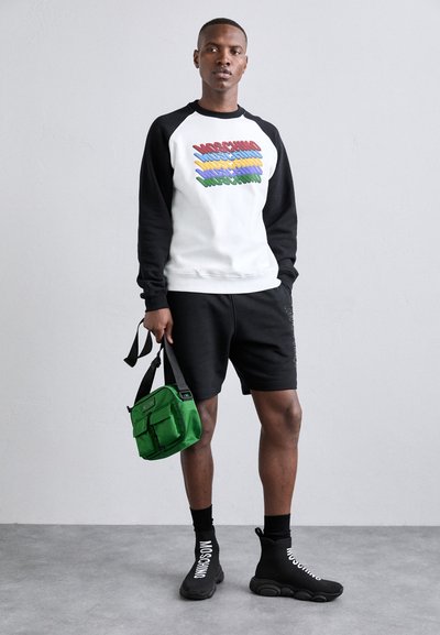 White sweatshirt with black sleeves, featuring colorful text. Black shorts, black socks, and black sneakers. Green crossbody bag with pockets.