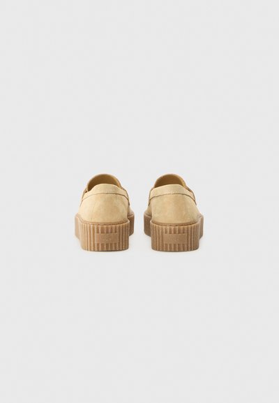 Chaussures slip-on en suède beige avec une semelle nervurée, dotées d'un bout arrondi et d'un dos lisse sans lacets ni fermetures.