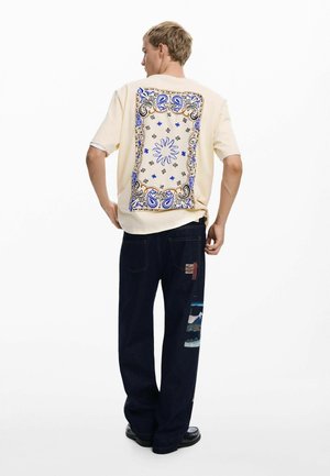 Cream short-sleeve shirt featuring a blue and gold paisley design on the back; paired with dark blue jeans with multi-textured patches.