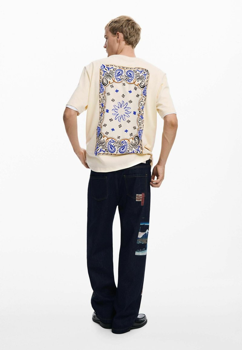 Cream short-sleeve shirt featuring a blue and gold paisley design on the back; paired with dark blue jeans with multi-textured patches.