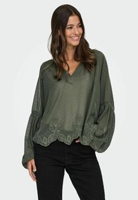 Donna che sorride, indossando una blusa verde oliva larga con ricami floreali e maniche a sbuffo, abbinata a pantaloni neri, su uno sfondo neutro.