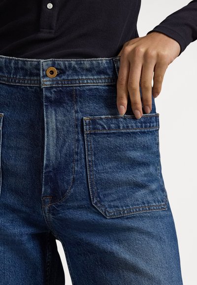 Άτομο που φοράει σκούρο μπλε τζιν με μεγάλα τετράγωνα μπροστινά τσέπες και μαύρο μακρυμάνικο πουκάμισο, το χέρι ακουμπισμένο στην τσέπη.