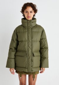 Doudoune vert olive avec un col haut, fermeture éclair, deux poches avant et une silhouette carrée, présentant une texture lisse et matelassée.