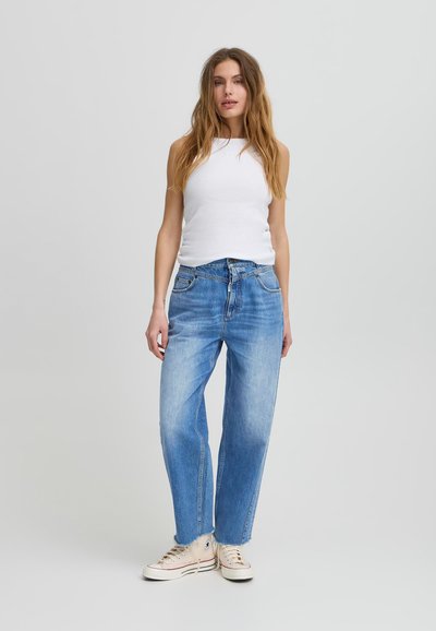 Gaiši zili platleņķa džinsi ar izbalējušiem akcentiem un augstu jostasvietu, apvienoti ar pielāgotu baltu bezdelīgu un bēšiem augstiem sporta apaviem.