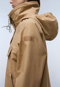 Osoba ubrana w beżową, wodoodporną kurtkę z kapturem, wysokim kołnierzem, regulowanym ściągaczem, kieszenią z klapą z przodu oraz naszywką z logo na górnej części rękawa.