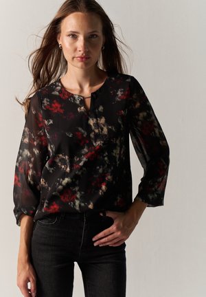 Mujer con cabello largo, vestida con blusa negra con flores y vaqueros negros, de pie con una mano en la cadera sobre un fondo liso.