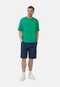 Groene katoenen t-shirt met korte mouwen, voorzien van een oranje en zwart grafisch ontwerp, gecombineerd met donkere denim shorts en witte sneakers.