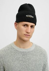 Sort strikket beanie-hat med ribbet tekstur og broderet "STUDIO" logo i hvid og guld. Grå, struktureret sweater med rund halsudskæring synlig.