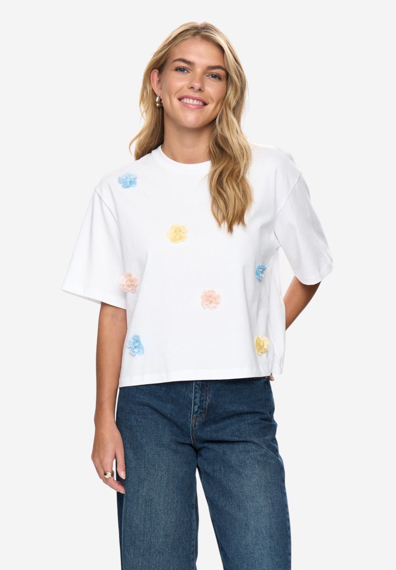 Hymyilevä nainen, jolla on pitkät vaaleat hiukset ja valkoinen T-paita, jossa on pastellinsinisiä, keltaisia ja vaaleanpunaisia kukkia, sekä siniset farkut.