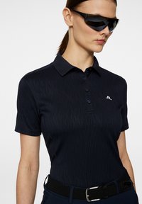 Marinblå pikétröja med ett strukturerat mönster, korta ärmar och krage. Har en liten logotyp på bröstet och ett svart vävt bälte.