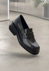 Černé kožené mokasíny s hladkým povrchem, nízkým zúženým podpatkem a dvojitými dekorativními pásky přes přední část. Minimalistický design, bez vzorů.