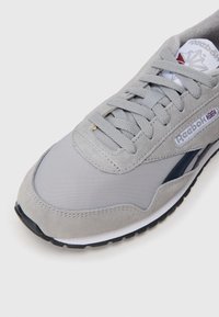 Sapatilha atlética cinza com uma mistura de materiais de camurça e rede, ponta redonda, sola robusta e um logo da Reebok na lateral.