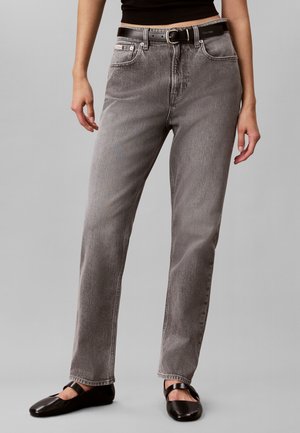 Person iført højtaljede grå jeans med sort bælte, sort kort top og sorte Mary Jane-stil sko mod en grå baggrund.