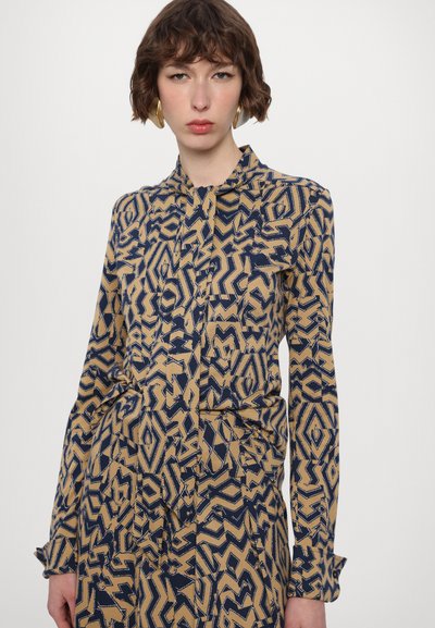 Long-sleeve shirt in tan with a bold navy abstract pattern. Features a button-down front, collar, and cuffed sleeves. Smooth texture.