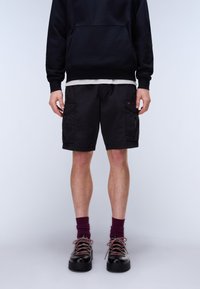 Uomo che indossa pantaloncini cargo neri, felpa nera con cappuccio sopra una maglietta bianca, calzini color vinaccia e scarpe di pelle nere con lacci viola, in piedi contro uno sfondo semplice.