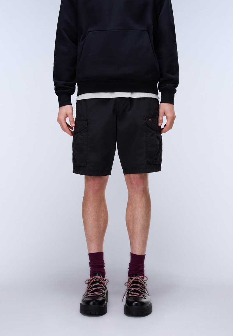 Uomo che indossa pantaloncini cargo neri, felpa nera con cappuccio sopra una maglietta bianca, calzini color vinaccia e scarpe di pelle nere con lacci viola, in piedi contro uno sfondo semplice.