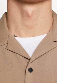 Beige overhemd met kraag en een gladde textuur, voorzien van een zwarte knoop en een wit ondershirt zichtbaar onder de kraag.