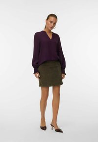 Donna in piedi con una camicia lunga viola, una gonna verde oliva e tacchi a punta marroni su uno sfondo bianco semplice.