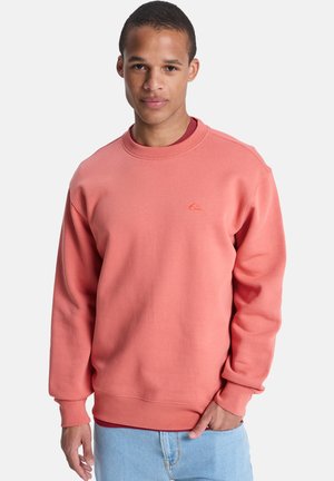 Koraalrode trui gemaakt van zachte stof, met een ronde hals, geribbelde boorden en een klein logo-accent op de borst. Gecombineerd met lichtblauwe spijkerbroek.