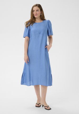 Kobieta w jasnoniebieskiej sukience midi z krótkimi bufkami i rozcięciami po bokach, stojąca z jedną ręką w kieszeni, w czarnych sandałach na obcasie.