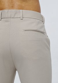 Zblízka na béžové kalhoty, které nosí osoba, zobrazení zadní kapsy a poutka na opasek na světlém pozadí.