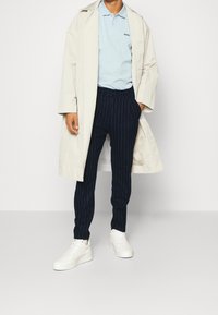En person som bär en beige trenchcoat, en ljusblå polotröja, marinblå randiga byxor och vita sneakers står mot en ljus enfärgad bakgrund.