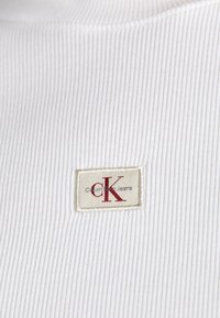 Valge sooniline kangas, millel on silt punaste "CK" tähtedega kreemitaustal, millel on kirjas "Calvin Klein Jeans." Sile tekstuur, lihtne disain.