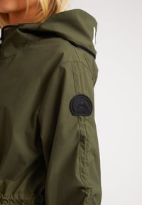 Kvinde iført olivengrøn jakke med hævet krave og sort rund "JACK1T" logo patch på den øverste ærme.