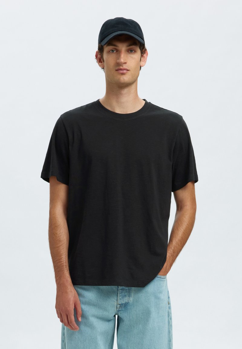 Joven de pie con una mano en el bolsillo, lleva gorra negra, camiseta negra y vaqueros azul claro sobre un fondo claro y liso.
