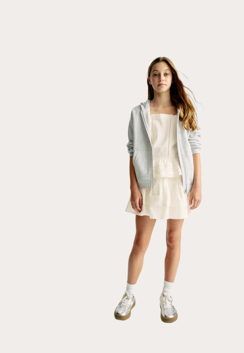 Ljusgrå zip-up hoodie över en vit lager-på-lager klänning. Silverfärgade sneakers med bruna sulor, vita ankelstrumpor. Enkel design med rena linjer.