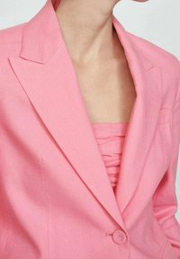 Blazer de lino rosa con solapa de pico, cierre de un solo botón y textura sutil. Debajo, se puede ver una blusa arrugada a juego.