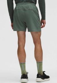 Mann in olivgrünen Sportshorts, passenden Lululemon-Socken, schwarzen Laufschuhen, steht mit entspannten Händen an den Seiten.