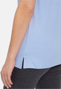 Světle modré tričko se bočním rozparkem na lemu, vyrobené z měkké látky. V páru s tmavě šedými legínami, které mají hladkou texturu.