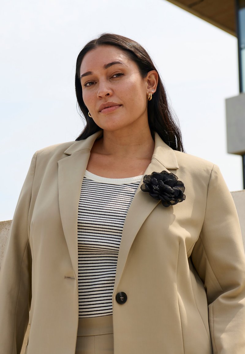 Blazer beige avec une épingle en forme de fleur noire sur le revers, porté sur un haut à rayures noir et blanc. Tissu lisse et coupe ajustée.