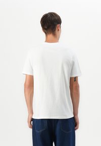 T-shirt en coton blanc avec un col rond et des manches courtes, vue de dos. Texture lisse, associée à un pantalon en denim bleu foncé.