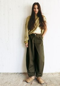 Olijfgroen oversized trui, lichtgele crop top, donkergroene wijde broek met ritsen, en beige veterschoenen met gevlochten accenten.