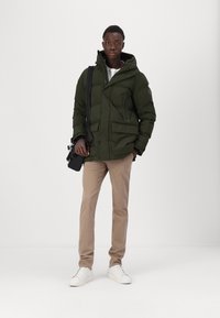 Grüne gepolsterte Jacke mit Kapuze, Fronttaschen und Reißverschluss; kombiniert mit beigen Hosen und weißen Sneakers. Schwarze Tasche über der Schulter.