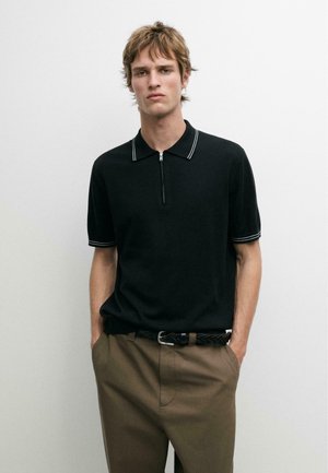 Giovane con capelli corti che indossa una polo nera a maniche corte con cerniera e pantaloni marroni con cintura intrecciata, mani nelle tasche, sfondo neutro.