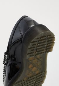 Zwarte glanzende zool van een nette schoen met een gestructureerd grippatroon en een gegraveerd logo, waarbij de hak en een deel van de schoenbovenkant worden weergegeven op een witte achtergrond.