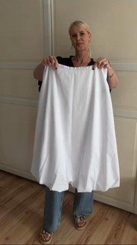 Kvinna med blont hår håller upp en stor vit kjol, stående inomhus på ett trägolv mot beige väggpaneler.