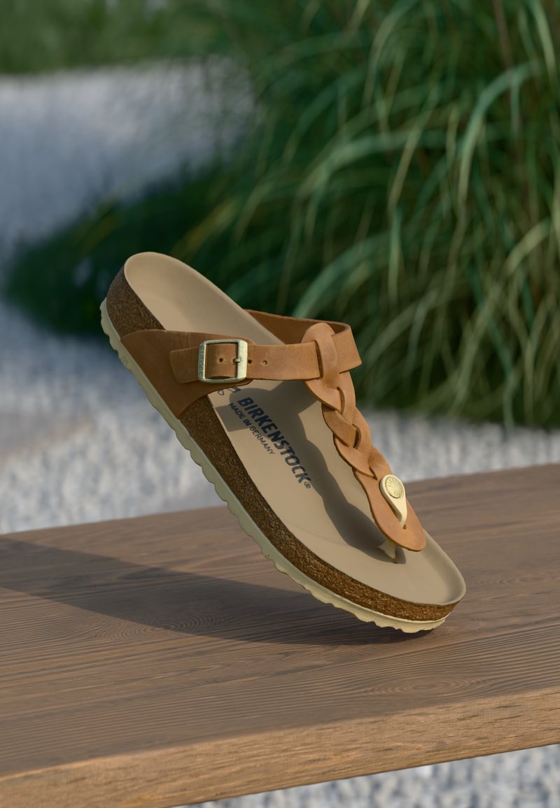 Ruda Birkenstock sandalo su sagtimi ir pynimu dirželiu guli ant medinės paviršiaus, apsupta švelnios žalios žolės ir žvyrų.