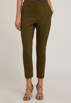 Kobieta w oliwkowo-zielonych, dopasowanych, krótkich spodniach i sandałach na wysokim obcasie ze wzorem wężowej skóry, stojąca z jedną ręką w kieszeni.