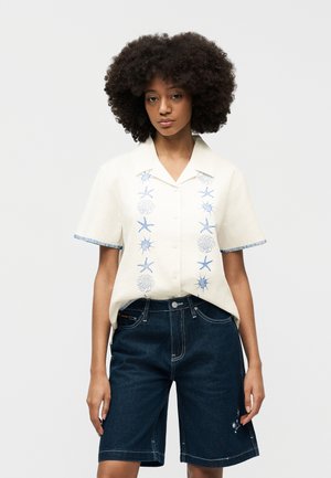 Afrofrizurás nő fehér, rövid ujjú blúzt visel kék tengerészmotívumos hímzéssel és sötét farmer térdnadrágot.