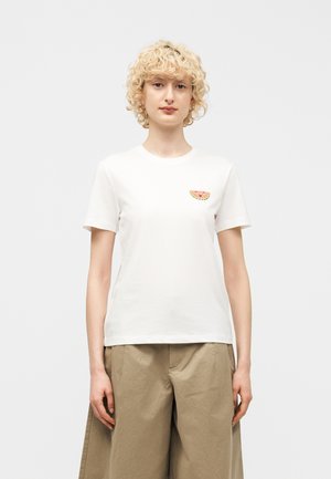 Ξανθό άτομο με σγουρά μαλλιά που φοράει λευκό μπλουζάκι με μικρό σχέδιο καρπουζιού και μπεζ παντελόνι φαρδιάς γραμμής, κοιτώντας μπροστά.