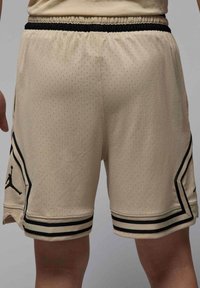 Pantalones cortos de baloncesto beige con una tela de malla texturizada, que presentan ribetes y acentos laterales negros, además de un logo Jumpman negro en la pierna izquierda.