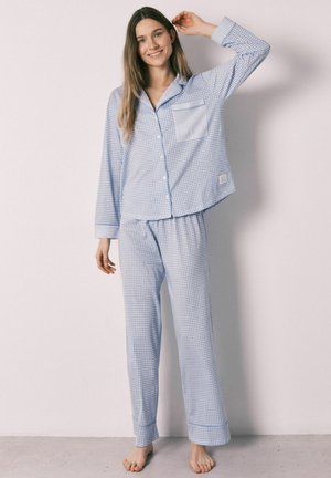 Ung kvinde, stående barfodet, smilende, iført lyseblåt og hvidt ternet pyjamassæt med lange ærmer mod en ensfarvet væg.