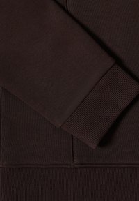 Zbliżenie na ciemno-brązową tkaninę sweatshirta, pokazujące mankiety, żeberkowy dół i detale szwu kieszeni.