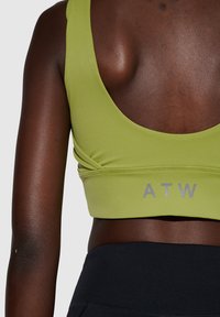 Grøn sports-BH med lav scoop ryg, lavet af glat stof. Har et gråt "ATW" logo ved bæltet. Sammen med sorte højtaljede leggings.