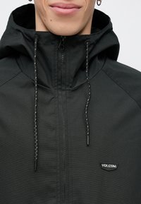 Veste noire à capuche avec fermeture éclair, en tissu texturé, cordons de serrage réglables et petit logo noir sur le côté gauche de la poitrine.
