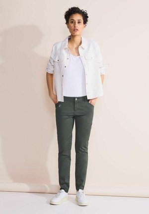 Bílá košile na knoflíky, bílý tílko, zelené slim-fit kalhoty, bílé tenisky. Pozadí je jemně růžové. Model stojí se s rukama v kapsách.
